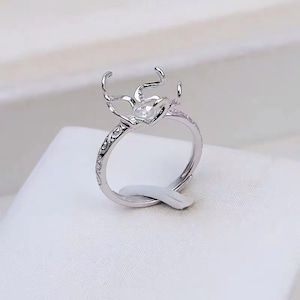 Peut inclure: Une bague argentée au design orné. La bague présente un sertissage en forme de cœur et des détails décoratifs le long de l'anneau. Le sertissage comporte plusieurs griffes s'étendant vers le haut. La bague est présentée sur une surface blanche.