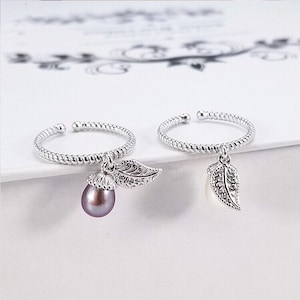 Könnte beinhalten: Zwei silberne Ringe mit einem baumelnden Blattanhänger. Ein Ring hat eine violette Perle am Anhänger.