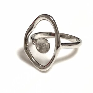 May include: A silver ring with an abstract design. The ring features a large, open, irregular oval shape with a small, round setting in the center. The band is smooth and simple. The ring is set against a white background.