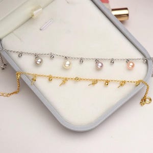 May include: Two delicate anklets. One is silver-toned with dangling pearls and small beads. The other is gold-toned, featuring small ankh symbols and beads. Both anklets are displayed inside a gray jewelry box.