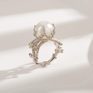 May include: A silver ring with a large white pearl set in a delicate, intricate design. The ring features a crown-like setting for the pearl, with branches extending from the base.