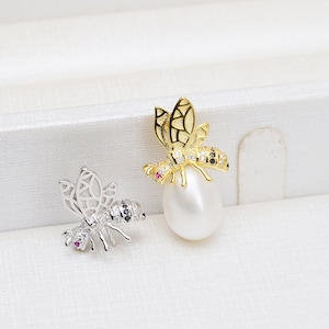 May include: Two bee-shaped pendants with black and white accents. One pendant is gold and the other is silver. Both pendants are on a white surface.