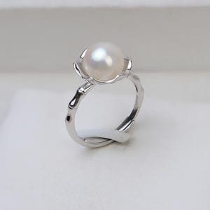 May include: A silver ring with a white pearl set in a flower-shaped design. The band of the ring is a simple, smooth silver.