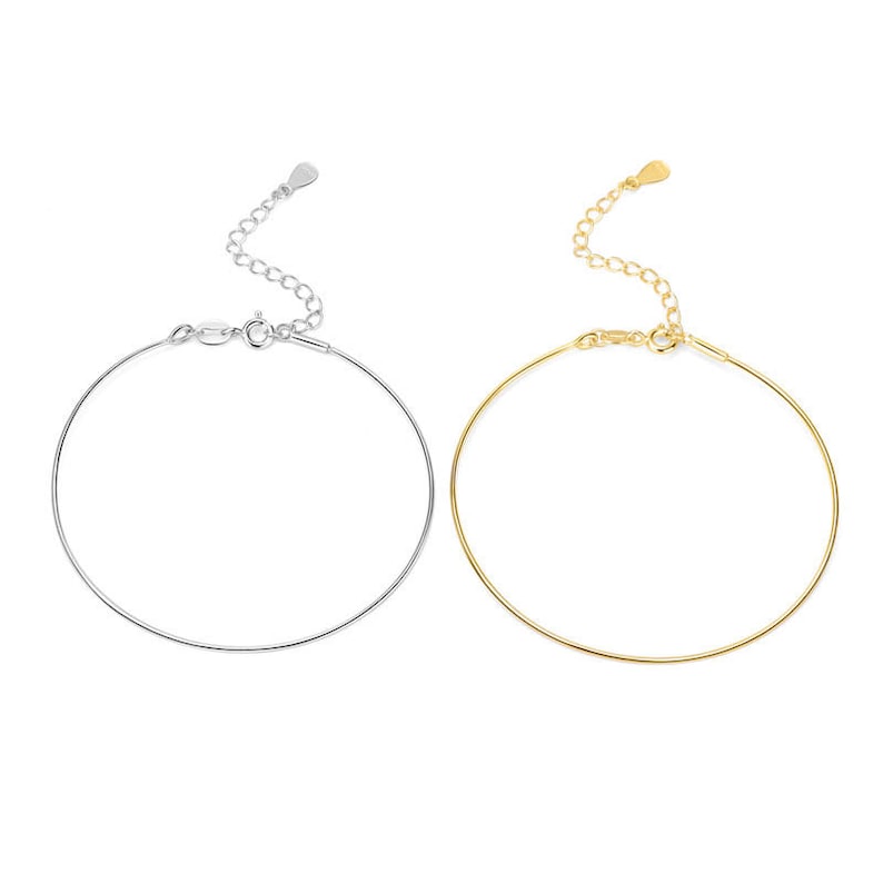 May include: Two delicate bracelets, one silver-toned and one gold-toned, are displayed against a white background. Each bracelet features a thin, circular band with an adjustable chain and clasp closure. The gold bracelet has a heart-shaped charm.