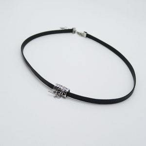 May include: A black choker necklace with a silver decorative charm. The charm is a cylinder with a floral design.