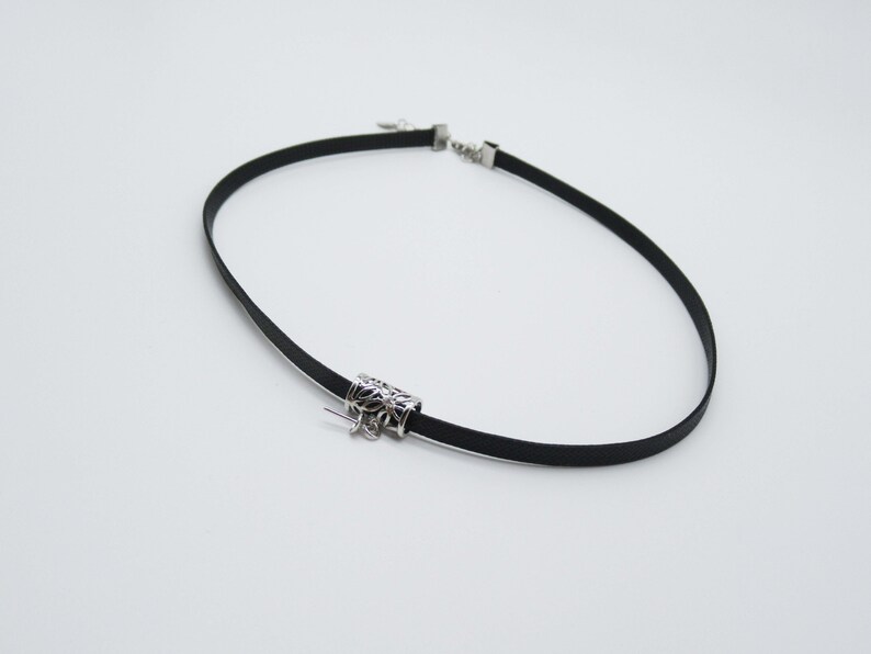 May include: A black choker necklace with a silver decorative charm. The charm is a cylinder with a floral design.