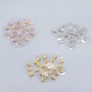 Puede incluir: Tres pilas de respaldos de pendientes, cada uno con un color de metal diferente: oro, plata y oro rosa. Los respaldos son de pl&aacute;stico transparente con un poste de metal.