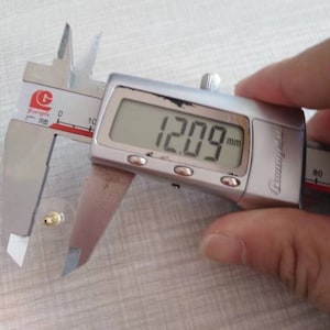 Puede incluir: Un calibre digital plateado que mide 12,09 mm. El calibre est&aacute; abierto y mide un objeto peque&ntilde;o de color dorado.