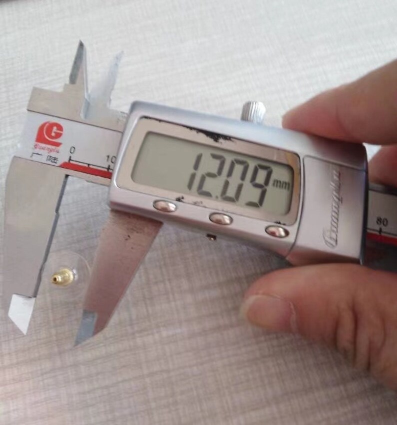 Puede incluir: Un calibre digital plateado que mide 12,09 mm. El calibre est&aacute; abierto y mide un objeto peque&ntilde;o de color dorado.