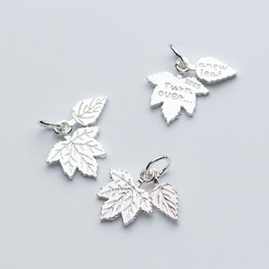 Könnte beinhalten: Drei silberne Anhänger in Form von Ahornblättern. Die Blätter haben eine strukturierte Oberfläche und das größere Blatt trägt die Gravur "It's a new leaf... says Turn over...".