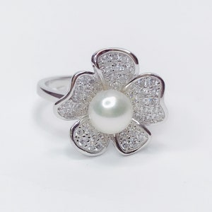May include: A silver ring with a flower design featuring a large pearl in the center and small clear stones around the edges.