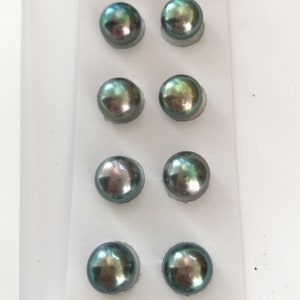 May include: Eight round, iridescent, dark green pearls in a white plastic packaging. The pearls are arranged in four rows of two.