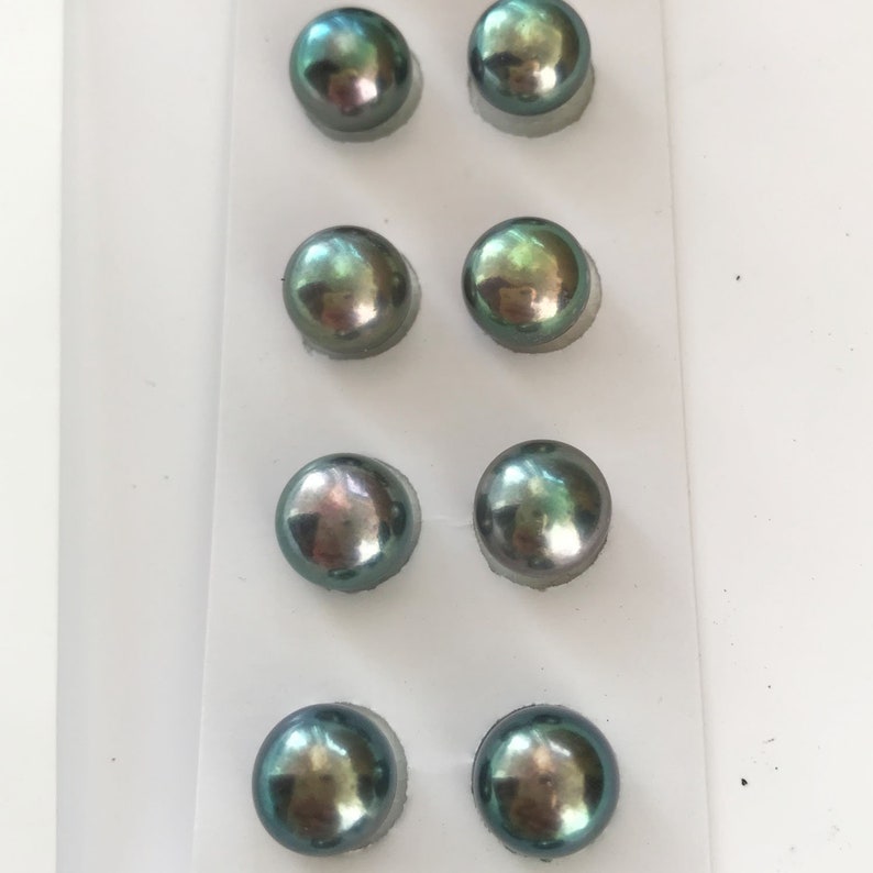 May include: Eight round, iridescent, dark green pearls in a white plastic packaging. The pearls are arranged in four rows of two.