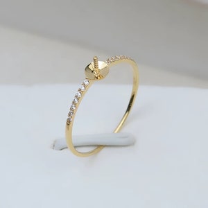 May include: A gold ring with a round setting and a band adorned with small, clear gemstones. The ring is set against a white background, highlighting its delicate design and craftsmanship. The setting is designed to hold a gemstone or pearl.