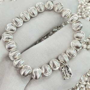 May include: A silver bracelet featuring textured beads, each with a unique pattern. The bracelet includes a small silver charm engraved with the words "You are the beloved sure".