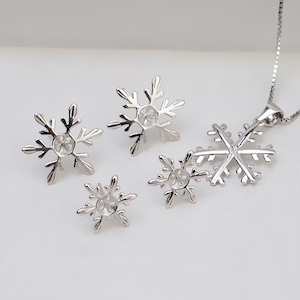 May include: A set of silver snowflake earrings and a matching pendant necklace. The earrings are in two sizes, and the pendant is larger. All three pieces feature a clear crystal in the center.