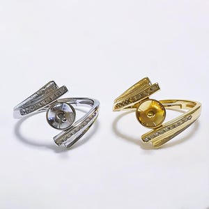 Può includere: Sono presentati due anelli, uno argentato e uno dorato. Ogni anello presenta un'incastonatura centrale ed è adornato da piccole pietre scintillanti. Le fasce si incurvano attorno al dito, creando un design moderno ed elegante. Gli anelli sono su sfondo bianco.