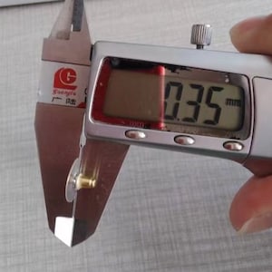 Puede incluir: Un calibre digital plateado que mide un peque&ntilde;o objeto de color dorado. El calibre muestra 0,35 mm.