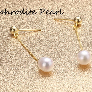 Puede incluir: Un par de pendientes de oro con una perla blanca que cuelga de cada pendiente. Los pendientes están sobre un fondo dorado. El texto "Aphrodite Pearl" es visible en la parte superior de la imagen.
