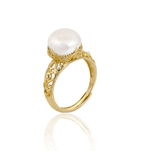 May include: A gold-colored ring featuring a large, round, white pearl. The ring has an ornate, filigree design on the band and around the pearl setting. The ring is displayed on a white surface.