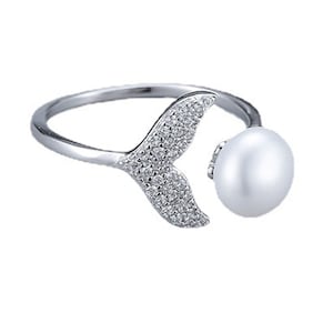 Puede incluir: Un anillo de plata con una perla y un diseño de cola de sirena. La cola está incrustada con pequeñas piedras transparentes.