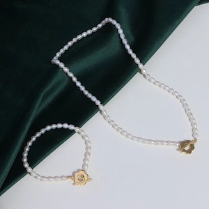 以下が含まれることがあります： 金色の花の形をした留め具が付いた真珠のネックレス2点セット。ネックレスは長く、ブレスレットは短いです。どちらも白い真珠で作られています。