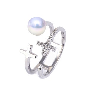 May include: A silver ring featuring two crosses, one adorned with small clear stones. A white pearl is attached to the top of the ring.