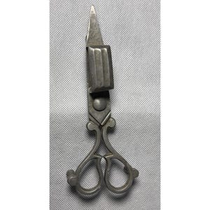 May include: A silver-toned metal wick trimmer featuring ornate, decorative handles with two finger loops and a rectangular box-shaped attachment on one blade designed to catch trimmed wick debris during the maintenance of candles.