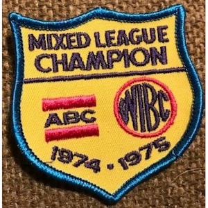 Puede incluir: Parche amarillo en forma de escudo con borde azul y la inscripción "MIXED LEAGUE CHAMPION" en morado. Presenta las letras "ABC" y "WBC" en rojo y morado, junto con los años "1974-1975".