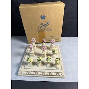 May include: A tic-tac-toe game set featuring a square board with a decorative border and nine individual game pieces. Each piece is a cream-colored pedestal topped with a cluster of pink or yellow flowers. The set includes a tan cardboard box labeled AVON Gift Collection.