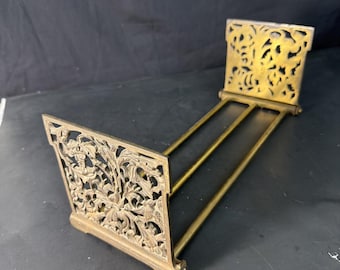 Puede incluir: Un sujetalibros de latón vintage con diseños florales y foliares calados y ornamentados. El sujetalibros tiene una base rectangular y un soporte trasero, conectados por tres varillas delgadas. El latón tiene una pátina cálida y envejecida.