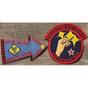 Op de afbeelding: Twee geborduurde patches. Eén is pijl-vormig, blauw met de tekst "Out of the Past" en "Into the Future", en een gele ruit met een wolf. De andere is cirkelvormig, marineblauw met een hand die naar sterren reikt, en de tekst "Reach for the Stars" en "Cub Express 1999".