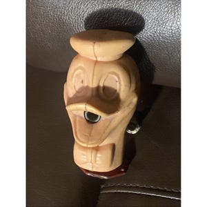May include: A vintage Donald Duck pencil sharpener. The pencil sharpener is made of a light tan plastic and features Donald Duck's face, hat, and bow tie. The sharpener has a small crank on the side and a red base.