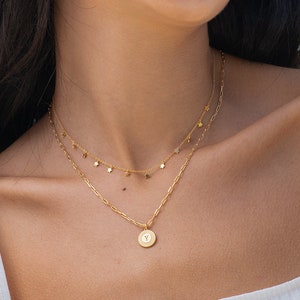 Puede incluir: Dos collares dorados. El collar superior tiene pequeños dijes colgantes. El collar inferior tiene un colgante circular con una letra. Los collares se usan sobre una camiseta sin mangas blanca.