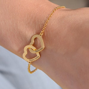 Op de afbeelding: Gouden kettingarmband met twee in elkaar grijpende hartcharms. De harten zijn gegraveerd met de namen "Betsy Sofia" en "Nicholas".
