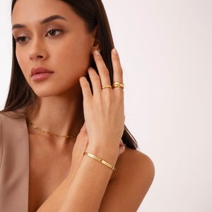 May include: A gold chain necklace with a delicate link design. The necklace is layered with a gold bangle bracelet with an engraved inscription. The bracelet is worn on the left wrist. The model is wearing two gold rings on her right hand.