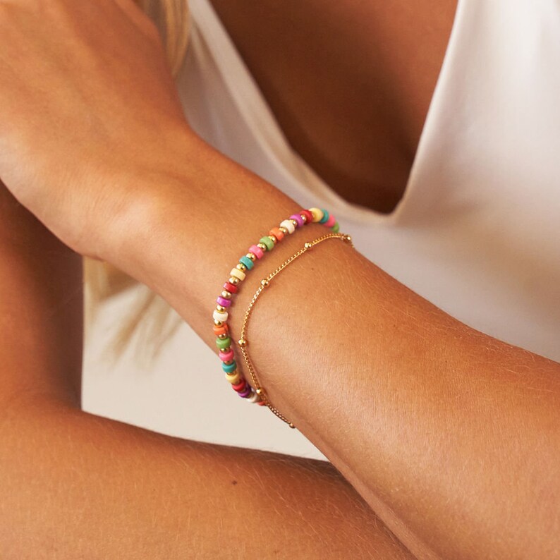 Pu&ograve; includere: Due braccialetti su un polso. Il braccialetto superiore &egrave; fatto di perline colorate e il braccialetto inferiore &egrave; una sottile catena d'oro.
