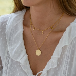 以下が含まれることがあります： 丸いペンダントとぶら下がっている文字が付いた、金色のレイヤードネックレス。 上の層には「LALA」の文字があり、下の層には小さなクリスタルが付いた円形のペンダントがあります。