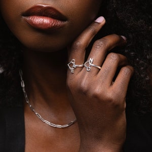 May include: Two silver rings spelling out "CAKE" and "NICE" are worn on a hand. A silver chain necklace is also visible. The rings have a delicate, modern, minimalist design. The image is a close-up, highlighting the jewellery.