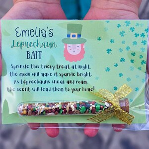 May include: A green card with the text "Emelia's Leprechaun Bait" and a cartoon leprechaun. The card is in a clear plastic bag with a small glass tube filled with colorful sprinkles tied with a gold ribbon.