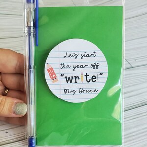 Puede incluir: Una tarjeta verde con una pegatina circular blanca que dice "Let's start the year off "write!" Mrs. Bruce". La pegatina tiene un dibujo de l&aacute;piz. Una bolsa de pl&aacute;stico transparente con un bol&iacute;grafo azul est&aacute; detr&aacute;s de la tarjeta.