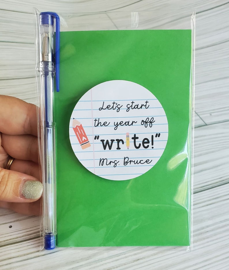 Puede incluir: Una tarjeta verde con una pegatina circular blanca que dice "Let's start the year off "write!" Mrs. Bruce". La pegatina tiene un dibujo de l&aacute;piz. Una bolsa de pl&aacute;stico transparente con un bol&iacute;grafo azul est&aacute; detr&aacute;s de la tarjeta.