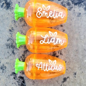 May include: Three orange carrot-shaped pencil sharpeners with green tops. Each sharpener has a name written on it: Emelia, Liam, and Atticus.