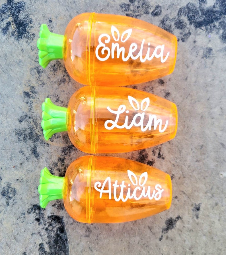 May include: Three orange carrot-shaped pencil sharpeners with green tops. Each sharpener has a name written on it: Emelia, Liam, and Atticus.
