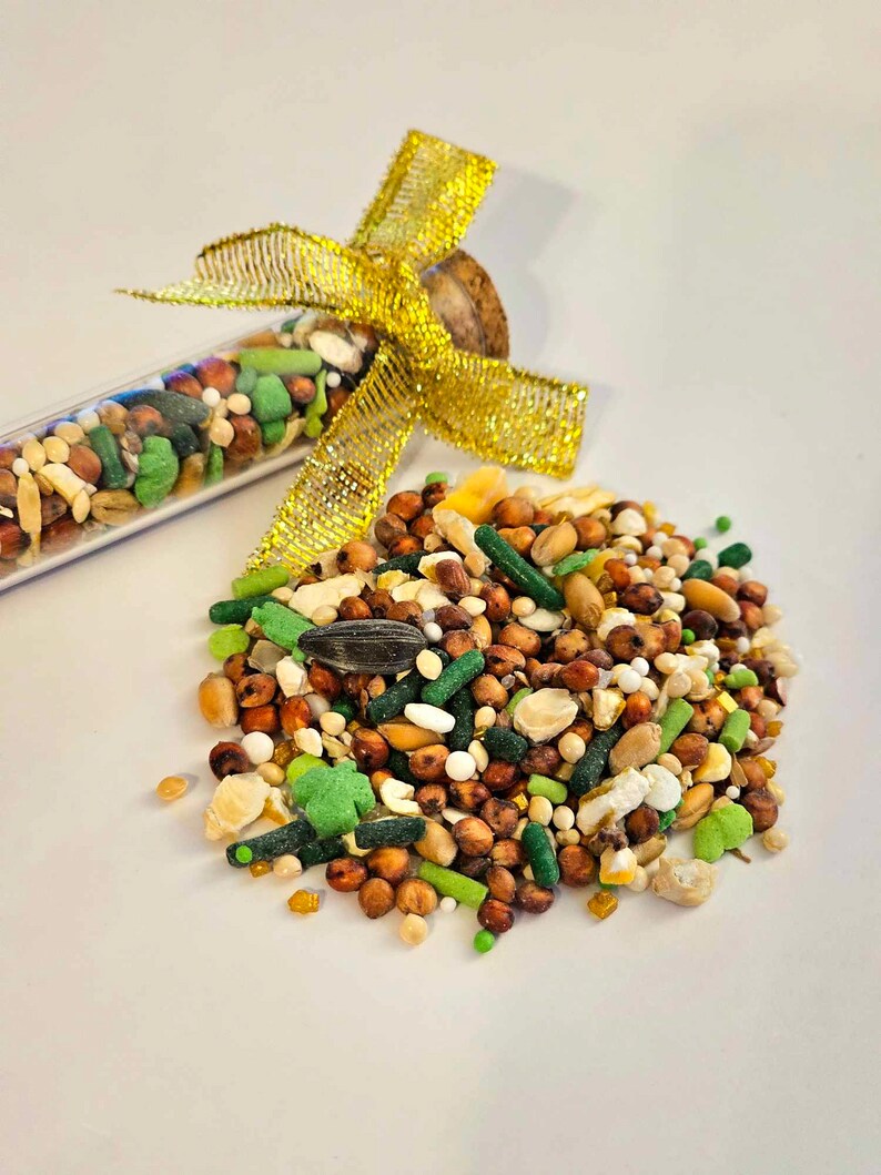 May include: A glass test tube filled with a variety of bird seed, tied with a gold ribbon. The tube is spilling out the seed onto a white surface.