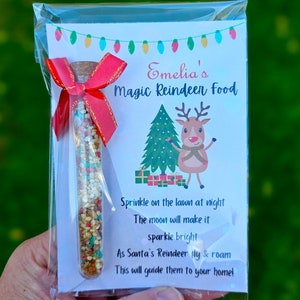May include: A small glass tube filled with colorful sprinkles, labeled "Emelia's Magic Reindeer Food". The label also includes a cartoon reindeer and a Christmas tree, with the text "Sprinkle on the lawn at night, The moon will make it sparkle bright, As Santa's Reindeer fly & roam, This will guide them to your home!"