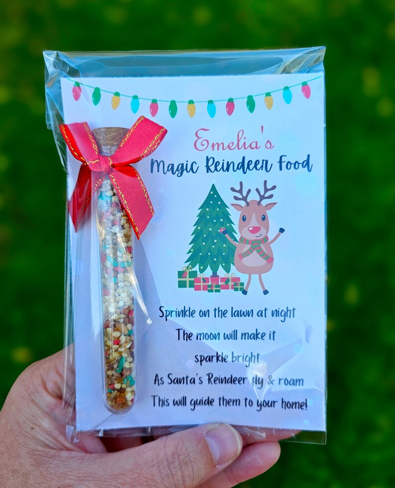 May include: A small glass tube filled with colorful sprinkles, labeled "Emelia's Magic Reindeer Food". The label also includes a cartoon reindeer and a Christmas tree, with the text "Sprinkle on the lawn at night, The moon will make it sparkle bright, As Santa's Reindeer fly & roam, This will guide them to your home!"