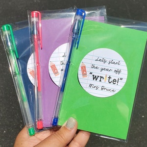 Puede incluir: Tres bolsas de pl&aacute;stico transparente que contienen cada una un peque&ntilde;o cuaderno y un bol&iacute;grafo transparente. Los cuadernos son morado, azul y verde. El cuaderno verde tiene una pegatina redonda en la parte delantera que dice "Let's start the year off "write!" Mrs. Bruce".