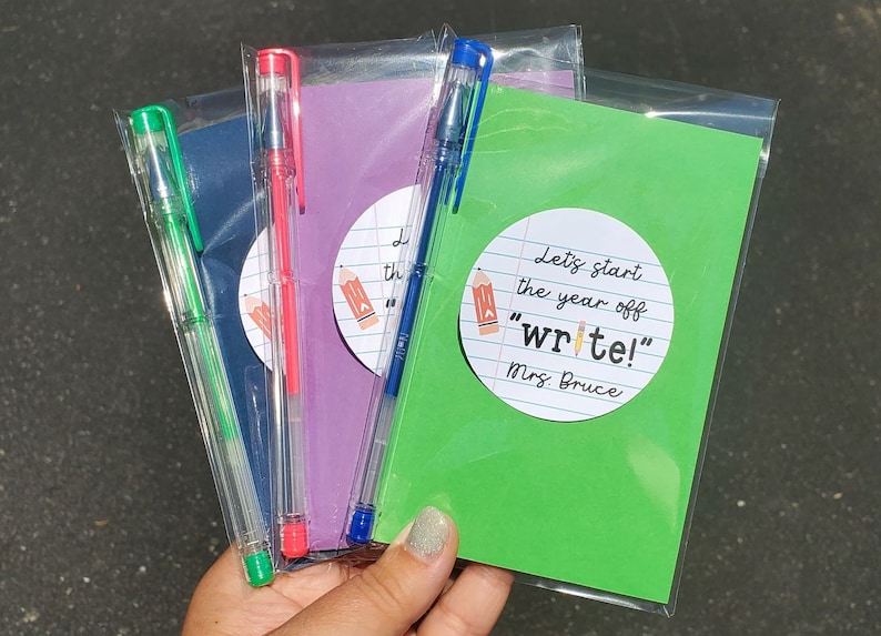 Puede incluir: Tres bolsas de pl&aacute;stico transparente que contienen cada una un peque&ntilde;o cuaderno y un bol&iacute;grafo transparente. Los cuadernos son morado, azul y verde. El cuaderno verde tiene una pegatina redonda en la parte delantera que dice "Let's start the year off "write!" Mrs. Bruce".