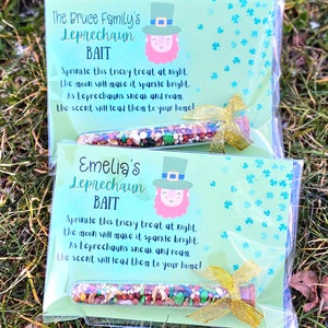 May include: Two clear plastic bags with green and white patterned paper inside. Each bag contains a small glass vial filled with colorful sprinkles and a gold ribbon. The paper inside each bag has a leprechaun illustration and the text "The Bruce Family's Leprechaun Bait" and "Emelia's Leprechaun Bait". The text reads: "Sprinkle this tricky treat at night. The moon will make it sparkle bright. As Leprechauns sneak and roam. The scent will lead them to your home!"